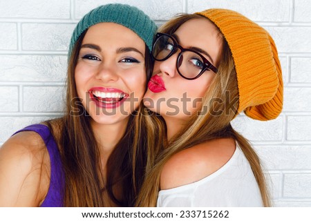 Close up fashion lifestyle portrait of two young hipster girls best friends, wearing bright make up and similar trendy hats, making funny faces and have gray time. Urban white brick wall background.