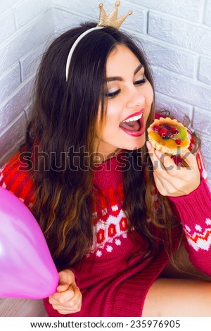 Young pretty girl having fun on birthday party, wearing party crown casual sweater, holding pink balloon and tasty small fruit cake. Positive emotions, read for celebration.