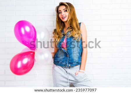 Young pretty stylish pretty blonde woman posing with big party balloons wearing hipster denim jacket and sportive grey pants, urban white background, bright colors.