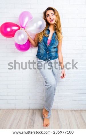 Young pretty stylish pretty blonde woman posing with big party balloons wearing hipster denim jacket and sportive grey pants, urban white background, bright colors, surprised emotions.