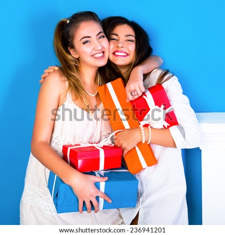 Indoor lifestyle portrait of two pretty young funny girls friends hugs smiling and having fun, holding bright holiday presents, ready for celebration. Wearing cozy pajamas.