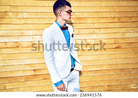 Fashion portrait of stylish man with hipster haircut, wearing trendy elegant white suit, denim shirt, bow tie and mirrored sunglasses, posing near wooden wall.  Street style look.