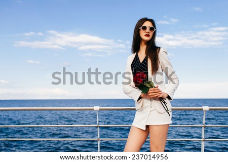 Outdoor fashion portrait of sexy brunette woman wearing trendy elegant white suit and sunglasses, holding red rouses, posing near sea an sunny day.