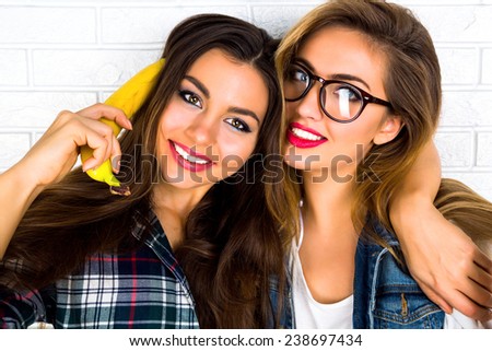 Lifestyle urban bright portrait of two young best friend girls going crazy and having fun together, playing with bananas imitating telephone, wearing glasses and bright sexy make up.