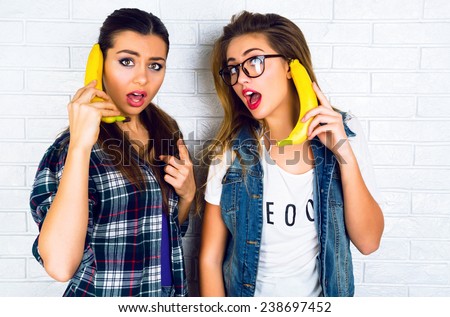 Lifestyle urban bright portrait of two young best friend girls going crazy and having fun together, playing with bananas imitating telephone, screaming and speaking with each other.