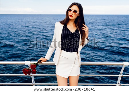 Fashion outdoor portrait of sexy beautiful woman with brunette hairs, wearing stylish white suit  and sunglasses, holding bouquet of red roses after date, amazing view on deep blue sea.