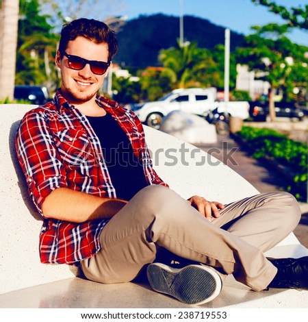 Outdoor lifestyle fashion image of young hipster stylish man sitting at the park wearing sunglasses plaid shirt and casual pants amazing view on mountains.