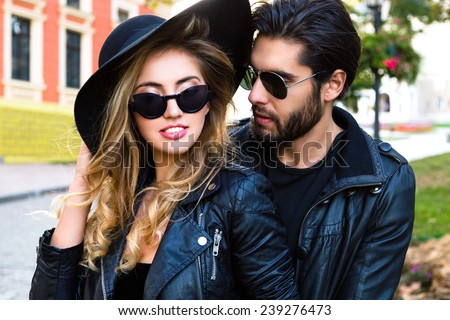 Close up outdoor fashion portrait of stylish young pretty couple in love hugs on the street of old town, wearing stylish total black leather rock n roll clothes ,sunglasses and hat.