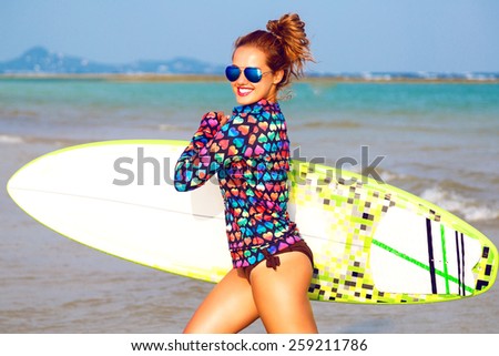 Outdoor summer portrait of sexy smiling woman running with surfer board near blue ocean,  wearing bright neon clothes and sunglasses, have fit tanned body and ginger hairs.