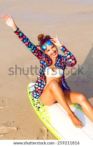 Outdoor fashion lifestyle summer portrait of stunning sexy surfer woman, wearing bright outfit makeup and sunglasses. Sitting on surf board smiling, put her hands in the air and enjoy her freedom.