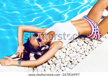 Young sexy woman laying and relaxed near pool at summer holiday nice hot day, wearing sexy mini shorts and crop top, listening and enjoy her favorite music at big headphones.