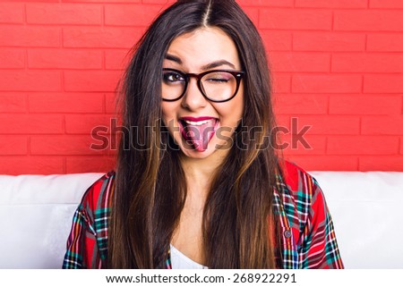 Fashion bright portrait of young woman with amazing long hairs and bright make up, having fun and showing long tongue at the room, wearing hipster outfit and glasses, red urban wall background.