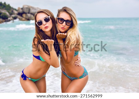 Two young brunette and blonde best friends looking on camera and sending you air kiss, have sexy slim body, wearing bikini sunglasses and fashion bright jewelry, posing in front of tropical beach.