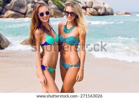 Two pretty best friends girls having fun on summer vacation, posing on amazing tropical beach with stones and clear blue water, wearing bright stylish bikini and sunglasses .