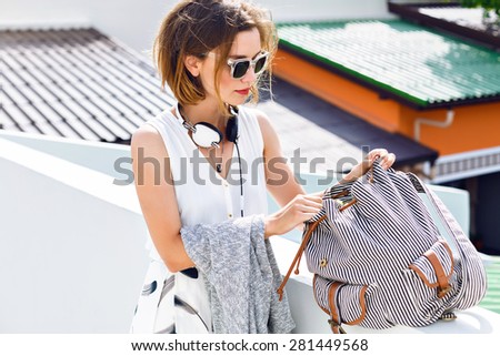 Close up fashion portrait of young hipster pretty woman, searching something at her backpack, walking and having fun at roof, stylish street style outfit.