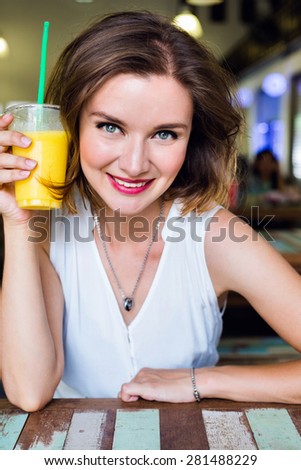 Indoor lifestyle fashion portrait of beautiful woman posing at cafe, drinking fresh healthy tasty mango juice, smiling, have nice time, bright sexy make up.