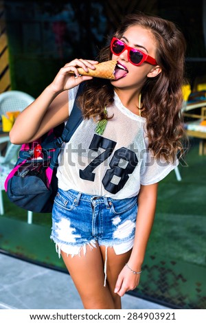 Funny beauty and sweet pleasure. An attractive young woman wearing sunglasses and trendy wear posing and eating ice cream on shoulder holding a gym bag in the summer outdoors.