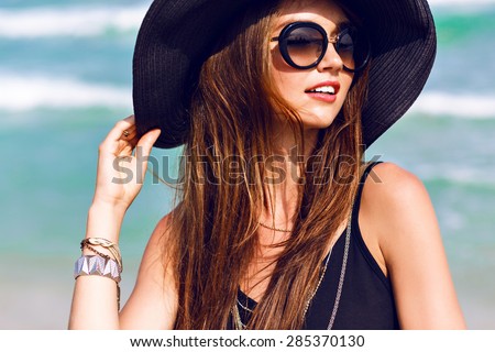 Close up sunny summer portrait of beautiful woman with fluffy brunette long hairs, smiling, having fun near blue ocean, wearing vintage sunglasses ,outfit and hat, vacation style , bright colors.