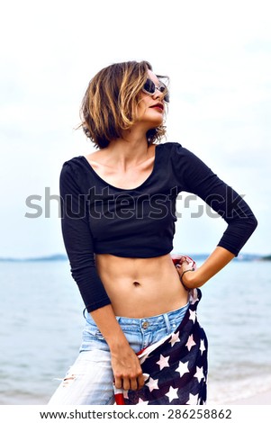 Summer fashion portrait of stunning fit sexy woman, wearing denim and crop top, holding american flag, spend time at the beach at rainy day.