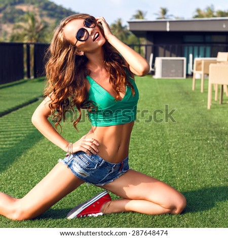 Summer portrait of sexy hipster woman, having fun on the roof at tropical island, wearing crop top denim micro shorts and sunglasses, brunette fluffy hairs, urban background. amazing smile.