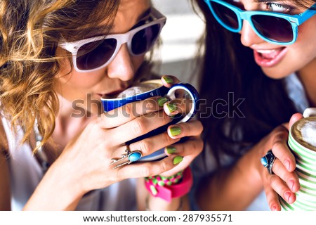 Close up lifestyle image of two young women drinking morning coffee speaking and gossip, bright stylish clothes sunglasses and accessorizes. two girls drinking cappuccino in cafe.