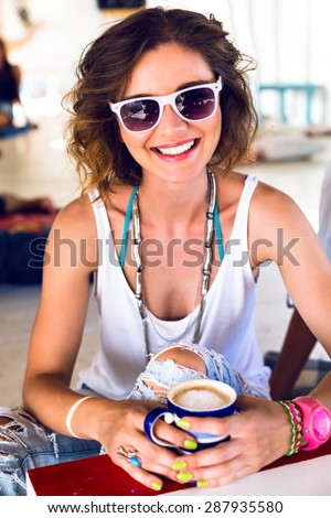 Summer sunny positive portrait of young pretty woman taking her morning coffee in cute cafe, wearing sunglasses and fashion jewelry, cute smile, joy, Saturday brunch.