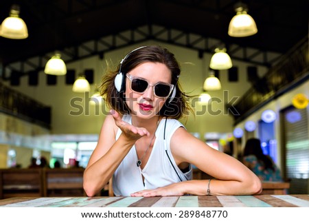 Young stylish pretty woman posing alone at city cafe, listening music, wearing trendy sunglasses and classic white shirt. sending kiss and smiling, joy, fun.