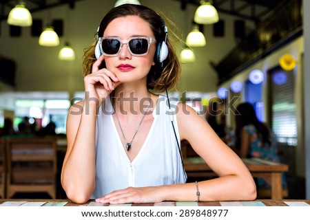 Young stylish pretty woman posing alone at city cafe, listening music, wearing trendy sunglasses and classic white shirt.