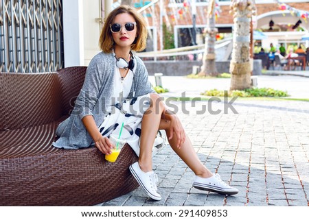 Outdoor lifestyle fashion portrait of woman posing on the street drinking orange smoothie, wearing stylish midi skirt, cardigan and sunglasses, joy, relax, vacation, travel.