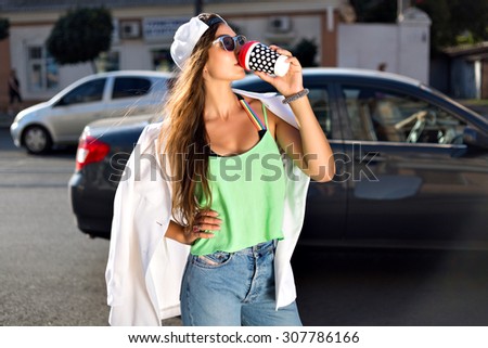 Lifestyle portrait of young hipster girl drinking take away latte on the street, wearing trendy summer vivid neon clothes hat and sunglasses, joy, travel, tasty drink.
