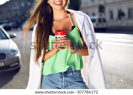 Lifestyle portrait of young hipster girl drinking take away latte on the street, wearing trendy summer vivid neon clothes hat and sunglasses, joy, travel, tasty drink. focus on hands.