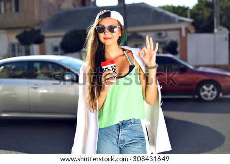 Lifestyle portrait of young hipster girl drinking take away latte on the street, wearing trendy summer vivid neon clothes hat and sunglasses, joy, travel, tasty drink, showing ok, by her hands.