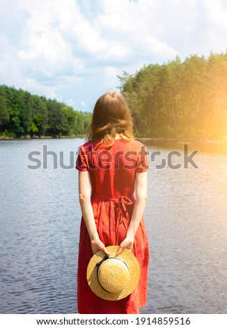 Beautiful girl with a straw hat near the lake. Amazing summer nature. Travel concept. Inspiration for wanderlust. Woman in linen red dress. Eco-friendly materials for clothes.