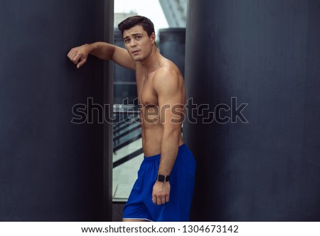 Portrait of calm tired bare-chested man with attractive body leaning on wall with hand while looking at camera