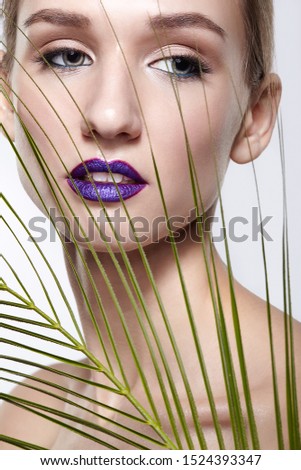 Female portrait with palm branch leaves on foreground. Woman has long neck and beauty face makeup with violet lips. Girl looking in camera with languid loving look.