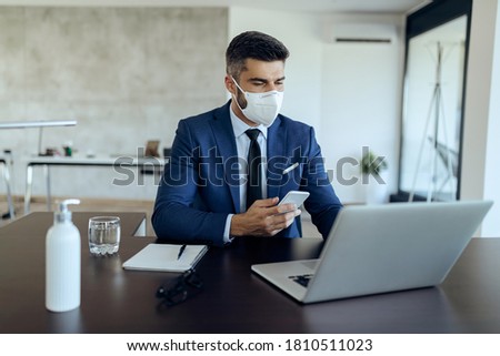 Male entrepreneur working on computer while using mobile phone and wearing face mask due to COVID-19 epidemic.  商業照片 © 
