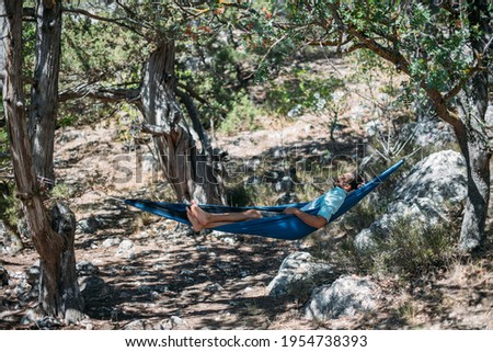 A man in a hammock on a hike in the mountains. Young handsome guy lies in a hammock in a camping in the mountains in the shade of trees and enjoys the surrounding nature on a sunny day