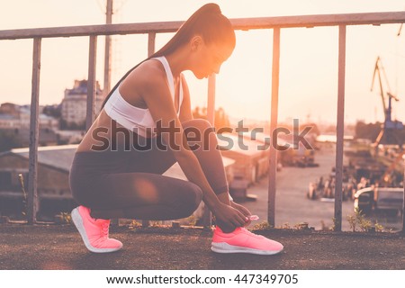 Tying her shoelaces. Beautiful young woman in sports clothing tying her shoelaces while standing on the bridge with evening sunlight and urban view in the background