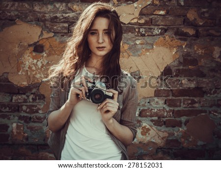 Young attractive female fashion model standing by the brick wall and holding retro camera.