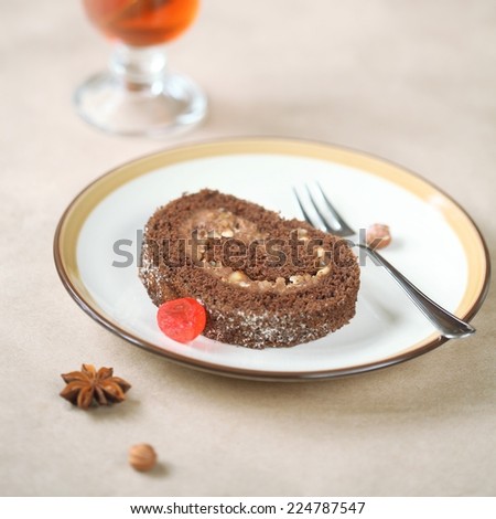 Piece of Chocolate Swiss Roll Cake with cherries and hazelnuts on a plate, silver fork and a cup of fruit tea, on a light beige background.