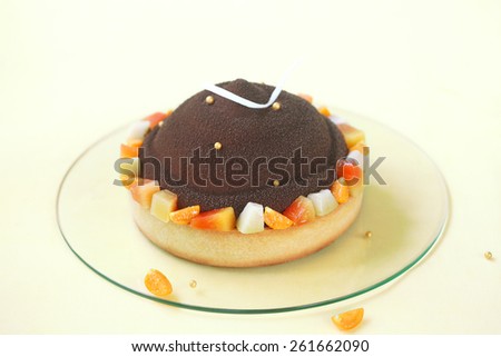 Chocolate, Mango and Macadamia Cake, decorated with tropical fruits and chocolate velvet spray, on a light yellow background.