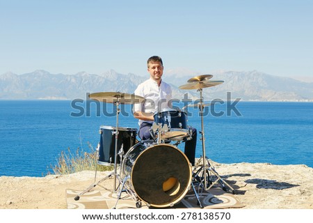 Young handsome guy in a white shirt drummer sitting behind the drum kit outdoors and unseat him the sea and beautiful sky