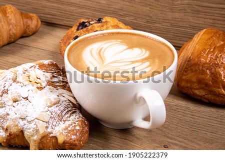 a white cup of cappuccino coffee with a heart shaped decorated foam with a selection of Danish pastry