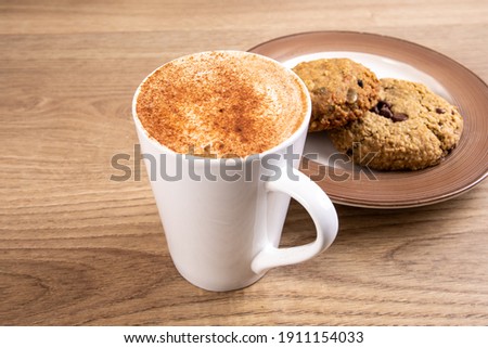 a white cup of chai latte coffee with cinnamon foam with a selection of cookies on a plate