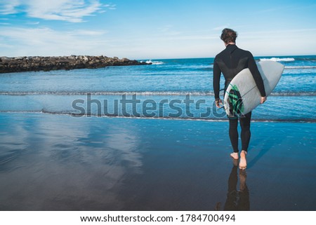 Young surfer entering into the water with his surfboard in a black surfing suit. Sport and water sport concept.