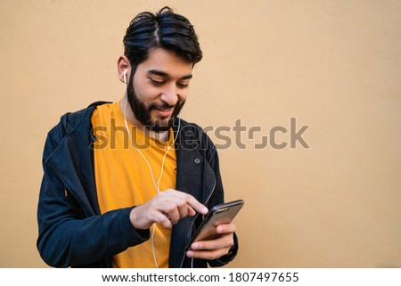 Portrait of young latin man using his mobile phone with earphones against yellow background. Communication concept.