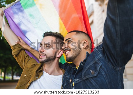 Portrait of young gay couple embracing and showing their love with rainbow flag at the street. LGBT and love concept.