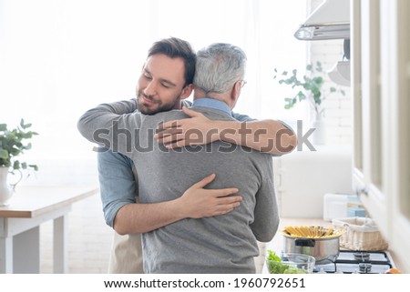 Cute loving caring old elderly senior father embracing hugging his adult caucasian son in the kitchen while cooking lunch, dinner, preparing meal together. Happy father`s day! I love you, dad!