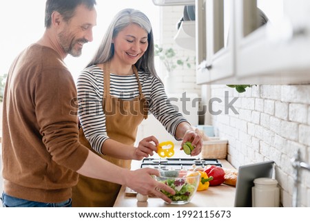 Happy mature middle-aged couple cooking vegetable vegetarian salad together in the kitchen, helping in preparation of food meal. Family moments, domestic homemade food