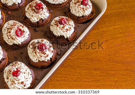many freshly baked chocolate muffins with whipped cream and cherries on top in a baking form on a wooden table